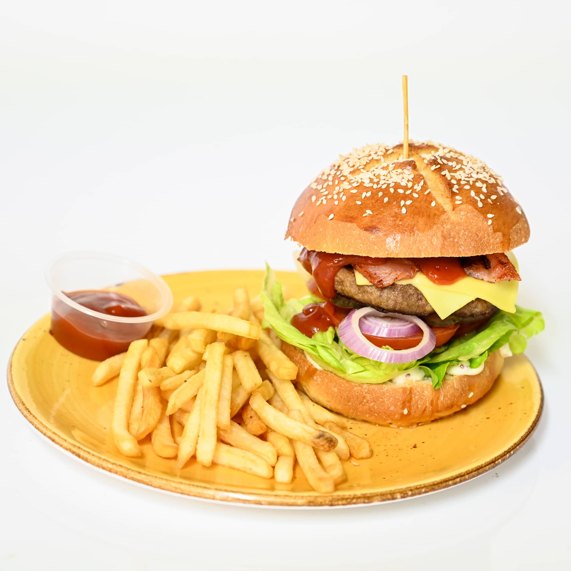 Hamburger with lettuce, tomato, and onion on a sesame seed bun with a side of French fries on a yellow plate.