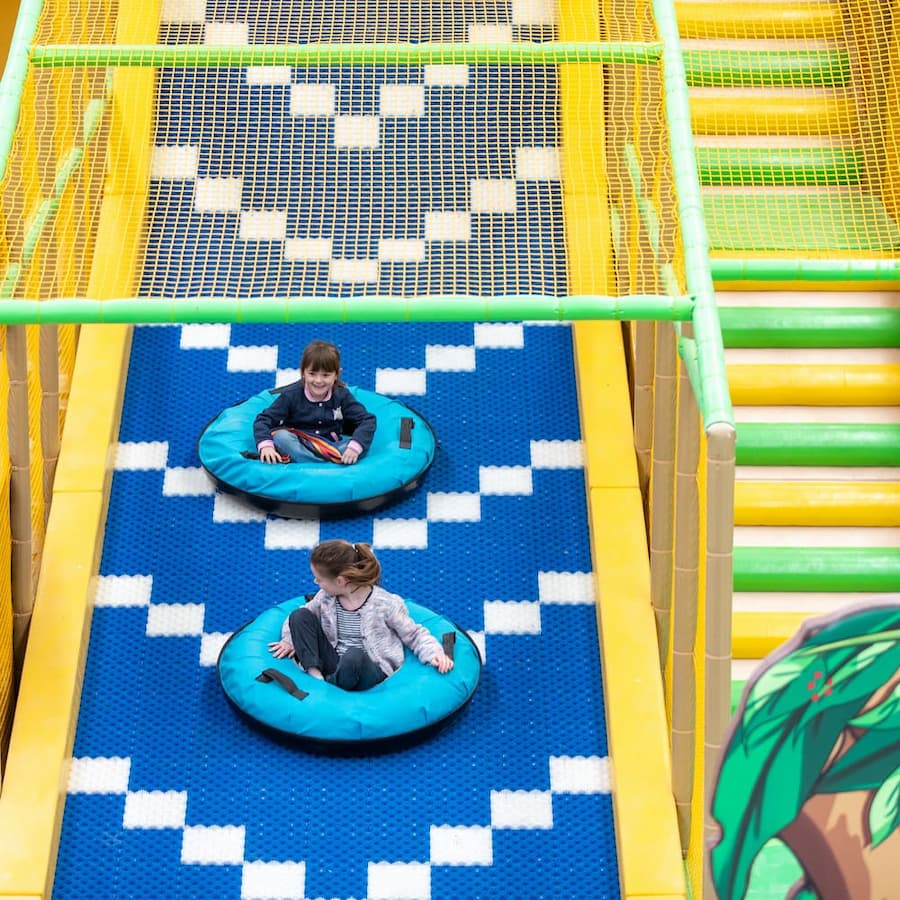 Two children sliding down a colorful indoor play structure on blue and white patterned mats, sitting in bright blue inner tubes. The play area has yellow and green safety netting and ladder-style steps.
