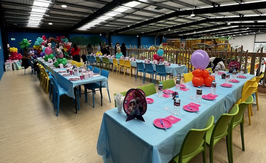 A large indoor event space with multiple long tables set up for a children's party. The tables are covered in light blue tablecloths, surrounded by colorful chairs in blue, yellow, and green.